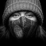 Black and white image of a person's face wearing a patterned fabric mask and a knitted beanie. The close-up photograph highlights their eyes, which are looking directly at the camera. The background is dark, emphasizing the subject.