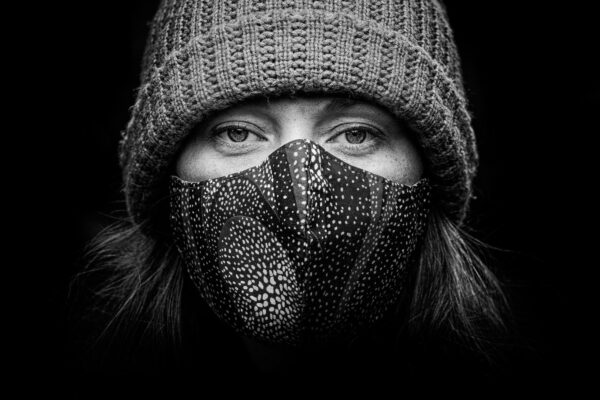 Black and white image of a person's face wearing a patterned fabric mask and a knitted beanie. The close-up photograph highlights their eyes, which are looking directly at the camera. The background is dark, emphasizing the subject.