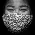 A close-up black and white photograph of a person's face wearing a leopard print face mask. The person's eyes are visible and they have an expression that appears neutral or slightly smiling. The background is solid black, emphasizing the subject.