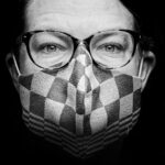 Black and white close-up of a person wearing glasses and a patterned face mask. Only the upper part of their face is visible, with light highlighting the details of their eyes and the texture of the mask. The background is dark, emphasizing the subject.