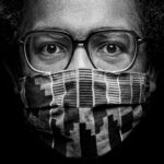 Black and white close-up photo of a person wearing glasses and a patterned face mask. The image focuses on their eyes which are looking directly at the camera, while the mask covers the nose and mouth. Their hair is curly and fills the sides of the frame.