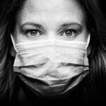 A black-and-white close-up photograph of a person with long hair, wearing a disposable surgical mask, covering the nose and mouth. The person has noticeable, expressive eyes looking directly at the camera, with a dark background enhancing focus on their face.