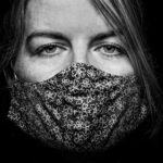 Close-up black and white photo of a person's face wearing a patterned cloth face mask. The person's eyes are looking directly at the camera, and their hair is partially covering one eye. The mask features a floral design. The background is dark.