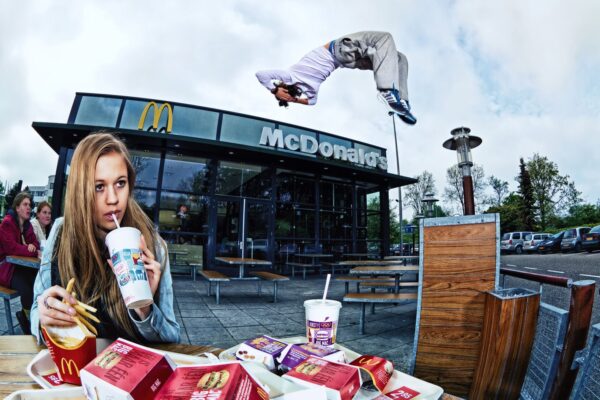 A young woman with long hair sips a drink while holding fries at a table outside McDonald's. Behind her, a person performs a high flip in mid-air. Several McDonald's food items are spread on the table. The restaurant's logo is visible in the background.