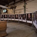 An indoor exhibition space displays a sequence of portraits hung on a fence, showcasing diverse individuals. The portraits are paired with written descriptions. The venue has a high ceiling with industrial structures, and large wooden crates are visible in the foreground.