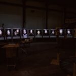 A dimly lit room displays a series of illuminated portraits on stands arranged in a semi-circle. The portraits show people's faces against dark backgrounds. The room contains other scattered objects, including a shopping cart and wooden structures.