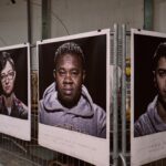 A series of large, framed black-and-white portraits of diverse individuals are displayed on metal fences in an indoor space. Each portrait features a person looking directly at the camera.