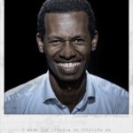 A man with a broad smile is centered in a portrait. He is wearing a light blue shirt, and a dark background highlights his face. The text reads: "Gizachew / Age: 28 / Ethiopia." Below, a quote says, "I wish for freedom in Ethiopia as in the Netherlands.