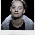 A close-up portrait of a woman with dark hair pulled back, wearing a black top and a gray fur-lined jacket. She has a neutral expression and looks directly at the camera. Text below reads, "Hadeel, Age 25, Syria. I wish to become a mother.