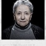 A grayscale portrait of an elderly woman with short gray hair, wearing a high-neck sweater and a black shawl. She stands against a dark background with a hopeful expression. Text below reads: "Suraja / Age: 69 / Azerbaijan. I wish for a good life in the Netherlands.