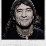 A man with long hair wearing a beanie, a necklace, and a dark shirt smiles slightly while looking directly at the camera. The caption below reads, "Usama, Age: 47, Libya. I wish to live like the Dutch people, very soon I wish!.