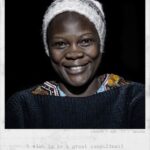 A smiling person wearing a white knit cap and a dark sweater with a patterned collar, facing the camera. The text below reads, "Zuenah / Age, 27 / Uganda. I wish to be a great consultant!.