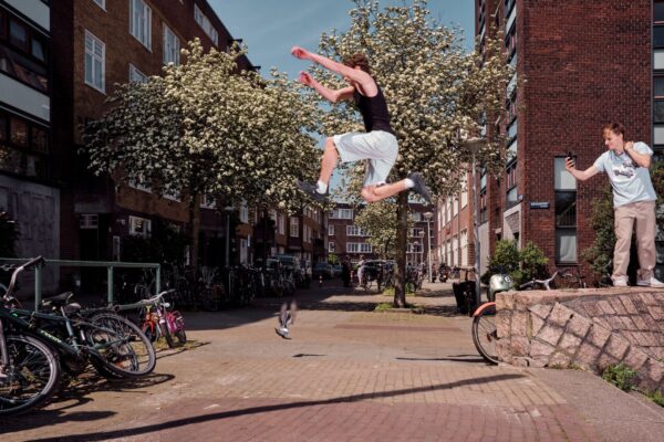 A person is mid-air performing a parkour jump between two areas in a residential neighborhood with blooming trees. Another person stands nearby, possibly cheering or recording the moment with a phone. Bicycles and buildings line the background on a sunny day.