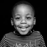 Black and white close-up photo of a smiling child wearing a striped shirt. The child has short hair and dimples, and the background is completely black.