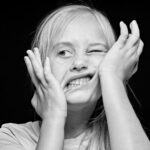 Black and white photo of a young girl with long blonde hair making a funny face by squishing her cheeks with her hands. She is wearing a short-sleeve shirt and has one eye partially closed while her mouth is twisted into an exaggerated expression.