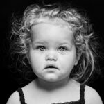 Black and white portrait of a young child with curly, tousled hair. The child has an inquisitive expression and is wearing a sleeveless top with delicate lace trim. The background is completely black, highlighting the child's face and features.