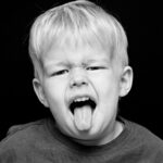 Black and white photo of a young child with light blonde hair, wearing a dark shirt, making a playful face by sticking out their tongue and squinting their eyes slightly. The background is solid black, highlighting the child's expressive gesture.