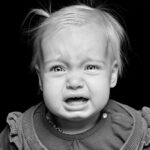 A black and white photo of a young child with light hair, looking distressed and crying. The child is wearing a long-sleeved top with ruffles on the shoulders. The background is completely black, making the child's expression stand out prominently.