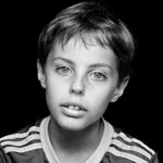 Black and white portrait of a young boy with short hair, wearing a striped shirt. The boy has a neutral expression and is looking directly at the camera against a solid black background.