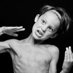 A young, shirtless boy with an expressive face and tousled hair wears a headband. He holds his hands up near his face, fingers spread wide, as if acting or making a playful pose. The background is black, emphasizing his animated expression and gesture.