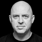 A black and white headshot of a bald middle-aged man with a neutral expression. He is looking directly at the camera against a dark background, wearing a plain dark shirt.