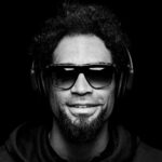 A black and white close-up portrait of a person with curly hair, wearing sunglasses, large over-ear headphones, and a hoodie. They are smiling and looking directly at the camera against a dark background.