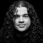 Black and white portrait of a person with long, curly hair, looking directly into the camera against a dark background. The expression is neutral and the lighting highlights the texture of the hair and face.