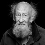 Black and white portrait of an elderly man with a long beard and wild hair. He is wearing a zip-up jacket and has a gentle smile. The background is dark, emphasizing his expressive face.