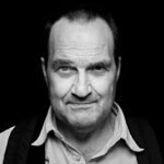 Black and white portrait of a middle-aged man with short hair and wearing a collared shirt and a backpack. He is looking directly at the camera with a neutral expression, set against a dark background.