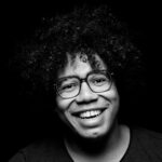 Black and white close-up of a person with curly hair and glasses smiling against a black background. They wear a dark shirt and their face is well lit with a joyful expression.