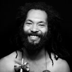 Black and white portrait of a smiling person with curly dreadlocks and a beard. The person has a joyful expression. They are shirtless, and a wooden object hangs around their neck. The background is dark, emphasizing their facial features.