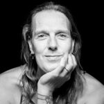 A black-and-white portrait of a person with shoulder-length hair and earrings, resting their chin on their hand while looking into the camera with a gentle smile. They have a tattoo on their hand and are wearing a tank top. The background is black, creating a stark contrast.