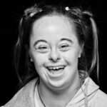 A black-and-white close-up photo of a person with Down syndrome, smiling widely. They have freckles and wear their hair in small pigtails, secured with hair ties. They are wearing a hooded jacket over a top, and the background is solid black.