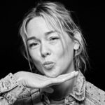 Black and white portrait of a woman with light hair styled in a loosely tied back manner. She is playfully posing with pursed lips, her chin resting on the back of one hand, and she is wearing a denim jacket adorned with small studs.