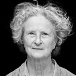 Black and white portrait of an elderly woman with curly, short hair and a gentle expression. She is wearing a collared shirt and facing the camera against a black background.