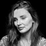 Black-and-white close-up portrait of a woman with long, wavy hair. She is gazing slightly downward with a subtle, contemplative expression. She is wearing a patterned blouse and layered necklaces, and the background is plain black.