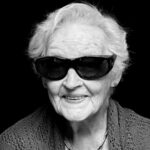 Black and white portrait of an elderly woman with short, wavy hair, wearing dark sunglasses and a textured cardigan. She is smiling gently at the camera against a black background.