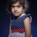 A young girl with dark, curly hair and a slight smile is posing against a dark backdrop. She is wearing a navy blue dress with white polka dots, a red ribbon around the waist, and floral patterns on the skirt. The lighting highlights her face and upper body.