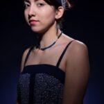 A young woman stands against a dark background, looking slightly to the side. She has dark hair tied back with a headband, wears a sleeveless patterned dress, and a beaded necklace. She has a calm, composed expression with a touch of red lipstick.