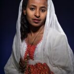 A woman is shown against a dark background, wearing a traditional white headscarf and a white garment with red and orange embroidery. She has dark hair and is looking directly at the camera with a calm expression.