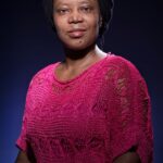 A woman stands against a dark background, wearing a bright pink, knitted top and a black head wrap. She appears confident, with a subtle smile and direct gaze. The lighting emphasizes her facial features and the texture of her clothing.