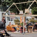 A group of people sits and stands under a wooden frame resembling a house outline, featuring cozy furniture like sofas and chairs, in an outdoor setting. Trucks are parked in the background, and there are trees and greenery around. A scooter is parked nearby.