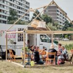 People sit and converse under a wooden structure resembling a house frame, set up outdoors near a white truck. The truck features various artwork and posters. A few buildings and greenery are visible in the background. Various items like chairs and a lamp decorate the area.