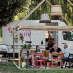 A group of four people sit at a table under a wooden gazebo with mismatched chairs and vintage lamps hanging above. Nearby, a baby stroller is parked. Behind them, a white truck is visible, parked under the shade of a large tree.