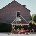 A group of people sit under a wooden frame resembling a house structure, placed on a quiet street next to a large brick house. They are relaxed on sofas and chairs, with a small table holding drinks nearby. The frame is decorated with string lights and a swing hangs from it.