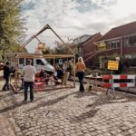 People gather under a wooden canopy structure on a cobblestone street. The area is adorned with a Dutch flag, string lights, and a yellow sign reading "Doorgang Verboden Afgezeten". Tables with items are around, and a van is parked nearby amidst a sunny sky.