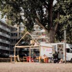 An outdoor scene features a white van parked next to a wooden frame resembling a small house. Several people, including children, are seated and standing around the setup under large trees. A multi-story residential building with balconies is visible in the background.