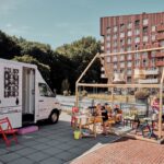 A mobile library truck is parked next to an outdoor reading corner with wooden tables and chairs under a pergola. Readers, including adults and children, are seated, enjoying books. A modern red-brick building and green trees are in the background.