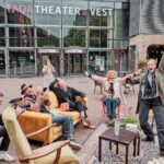 A group of people are joyfully gathered in an outdoor seating area in front of a building with "TAQA THEATER DE VEST" signage. Some are sitting on couches, while others are standing, all with exuberant expressions and hand gestures suggesting celebration.