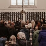 A crowded room in an art gallery with many people gathered around, observing a large wall display of black-and-white portraits. The space features high ceilings, white walls, and a chandelier hanging from the center of the ceiling.